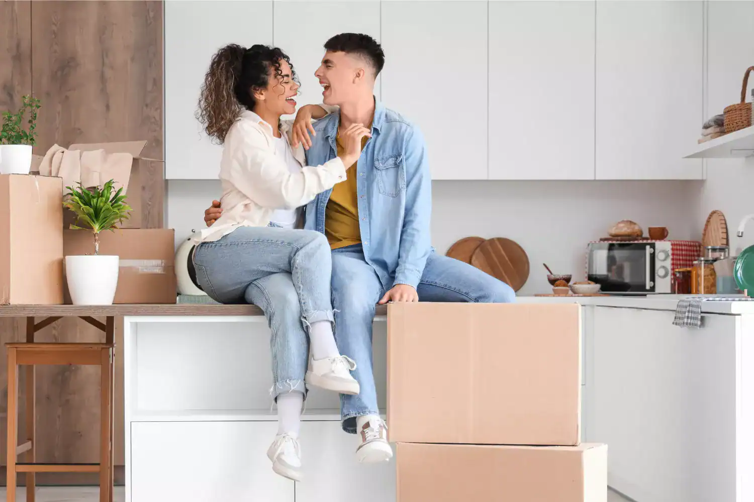 A young couple sitting together on a kitchen counter surrounded by moving boxes, unpacking and laughing as they settle into a new home while living together before marriage.