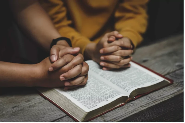 Two people sit closely with hands clasped in prayer over an open Bible on a wooden table.