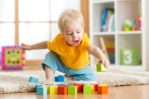 16-month-old toddler playing with colorful wooden blocks on the floor in a bright, cheerful room.
