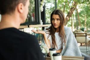 Insulted young woman gesturing with hand and looking at her boyfriend while sitting in a cafe at the park outdoors