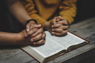 Two people sit closely with hands clasped in prayer over an open Bible on a wooden table.