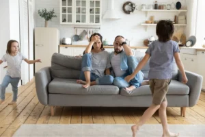 Exhausted parents sitting on a couch with their hands on their heads while their children run around, illustrating parental burnout.