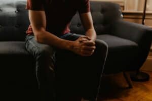 This image shows a man sitting on a couch with his hands clasped together in front of him, resting on his knees. What Does the Bible Say About Therapy?