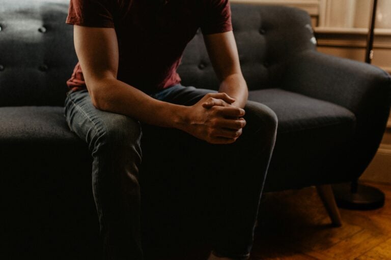 This image shows a man sitting on a couch with his hands clasped together in front of him, resting on his knees. What Does the Bible Say About Therapy?