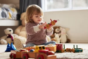 Toddler playing with wooden toy train while sitting on carpet surrounded by traditional baby toys