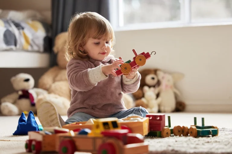 Toddler playing with wooden toy train while sitting on carpet surrounded by traditional baby toys