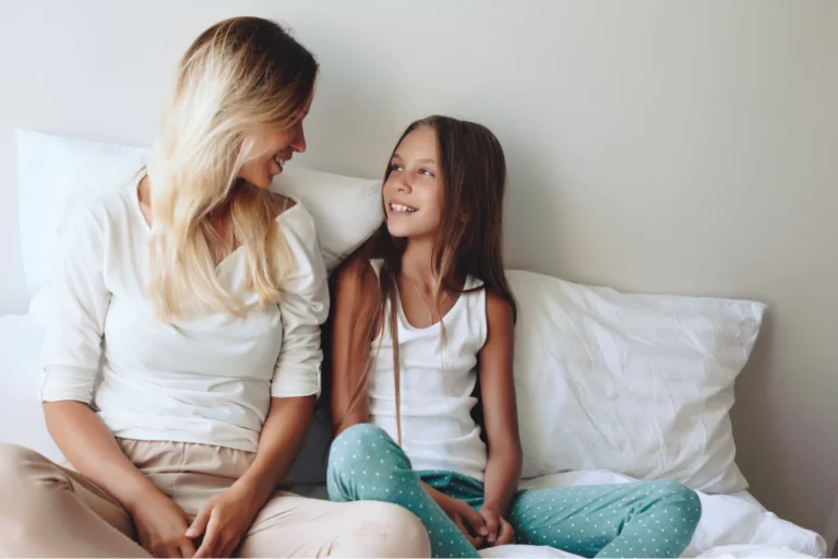 Mother and daughter chatting on a bed, smiling warmly, showing how to talk to 7–10 year olds in a safe, relaxed setting.
