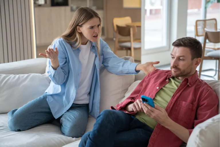 Woman yelling at spouse who ignores her while looking at phone on couch.
