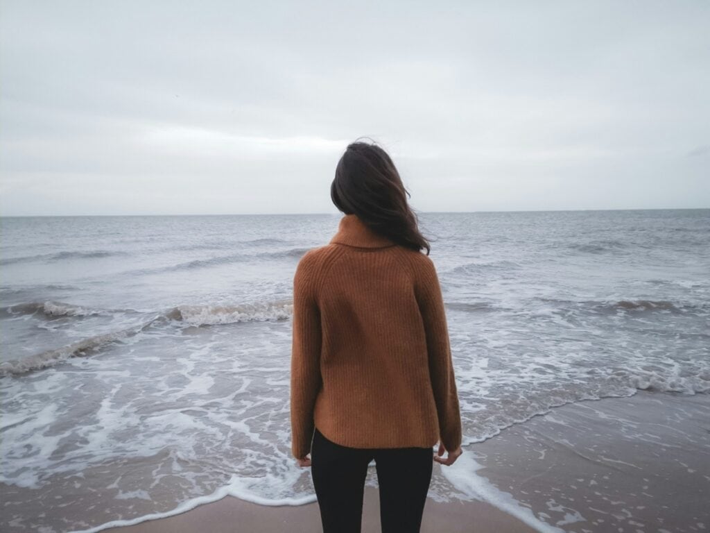 This image shows a woman standing at the edge of the water at a beach on a cloudy day. The waves are rolling in over her feet as she looks out towards the horizon. (Bible Verses About Anxiety)