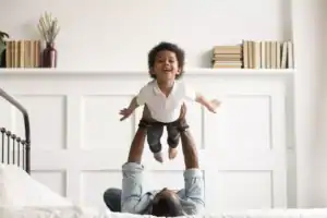 Father lifting his joyful son into the air while lying on a bed, capturing how dads parent differently than moms through playful bonding.