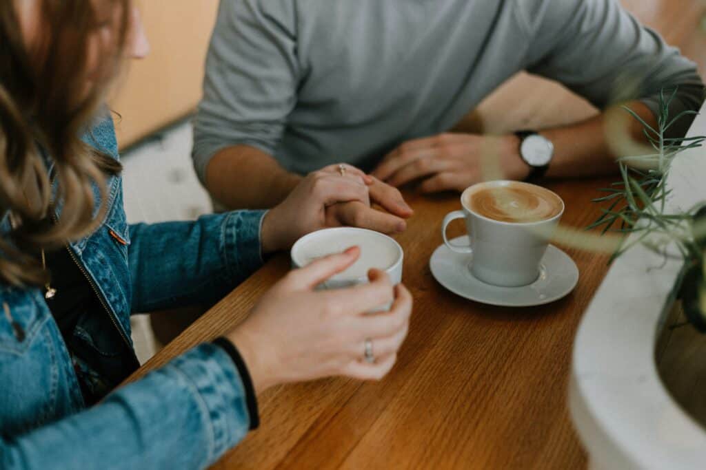 This image shows a couple sitting down at a coffee shop, holding hands with their mugs in front of them.