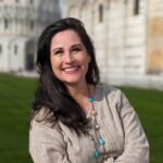 Jacqui Jackson smiling outdoors in front of a historic stone building, wearing a beige sweater and a necklace with turquoise accents.