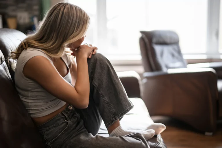 Teen girl sits curled up on a couch in deep thought, illustrating the emotional weight of mental health issues.