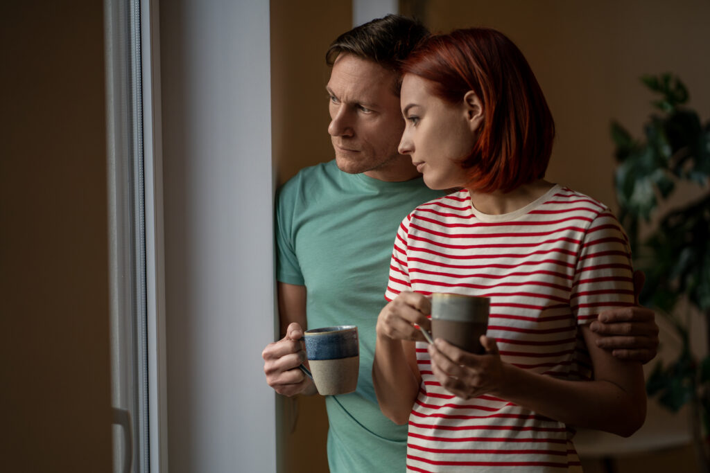 Man and woman with mugs of coffee look out window. Husband wife with hot cups of tea watching what happening outdoors, assessing cloudy rainy cold weather. Reluctance to leave house, day off, weekend