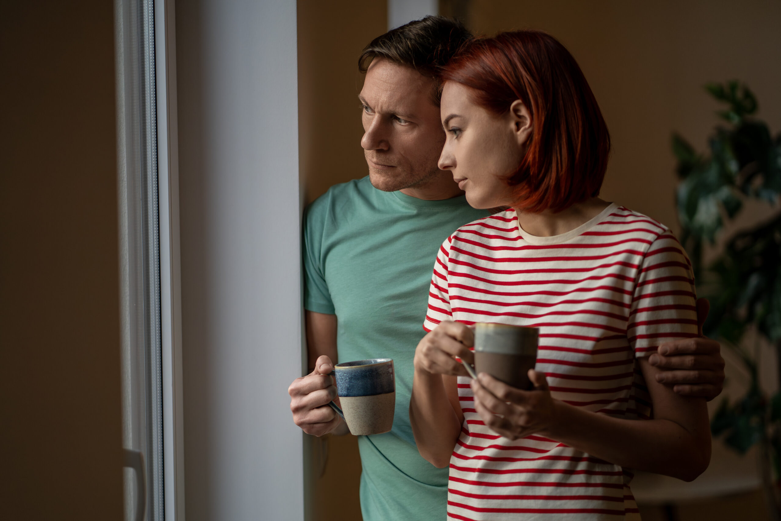 Man and woman with mugs of coffee look out window. Husband wife with hot cups of tea watching what happening outdoors, assessing cloudy rainy cold weather. Reluctance to leave house, day off, weekend
