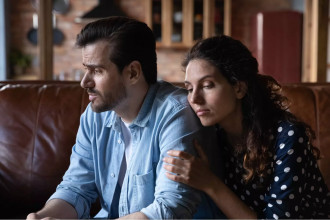 A couple sits close together on a couch, looking distressed and withdrawn, symbolizing the emotional process of healing from infidelity.