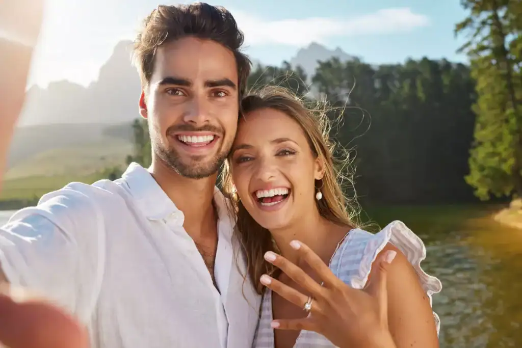 Couple celebrating their engagement outdoors, showing an engagement ring while taking a selfie, illustrating themes related to finding the best age to marry.
