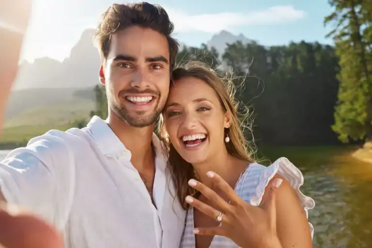 Couple celebrating their engagement outdoors, showing an engagement ring while taking a selfie, illustrating themes related to finding the best age to marry.