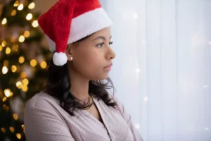 Young woman wearing a red Santa hat and light-colored blouse sitting indoors near a softly lit Christmas tree with warm golden lights.
