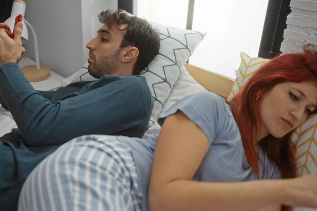 Couple lying in bed facing opposite directions, each holding a smartphone, symbolizing emotional distance and disconnection in marriage.