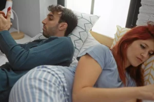 Couple lying in bed facing opposite directions, each holding a smartphone, symbolizing emotional distance and disconnection in marriage.