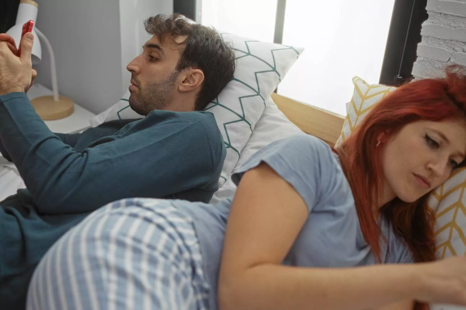 Couple lying in bed facing opposite directions, each holding a smartphone, symbolizing emotional distance and disconnection in marriage.