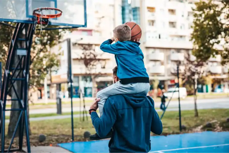 A father lifts his young son on his shoulders as the boy practices shooting a basketball, illustrating biblical masculinity through guidance, encouragement, and nurturing connection.