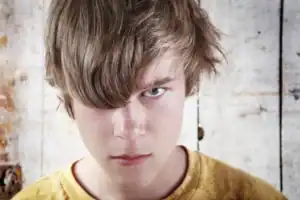 Teen with light brown, tousled hair standing in front of a weathered wooden wall.