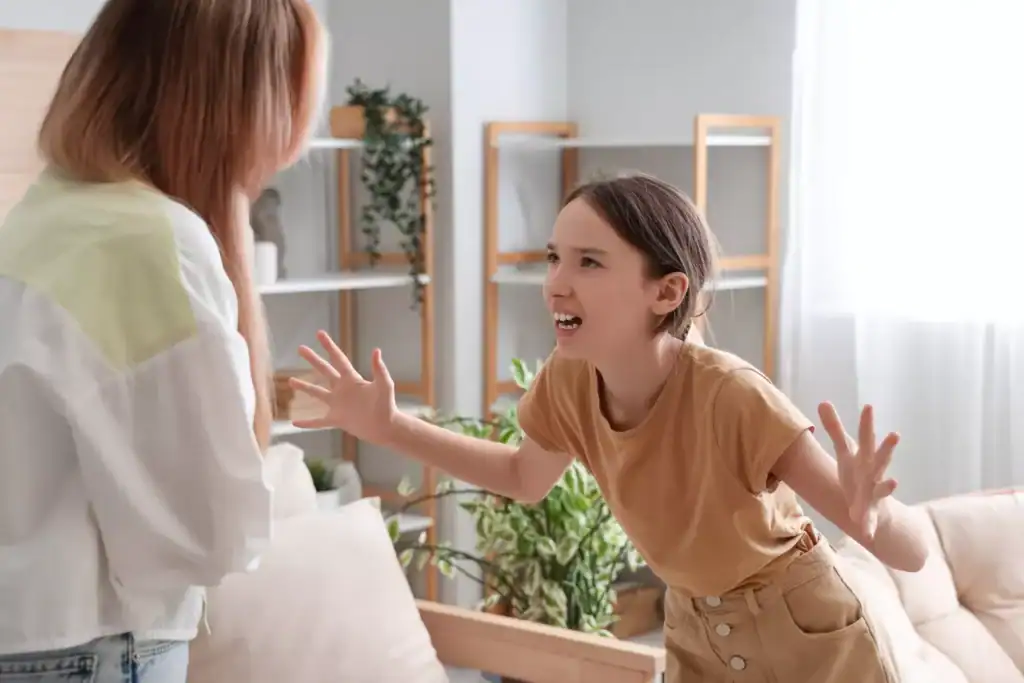 A little girl stands in front of her mom in a living room, raising both hands while arguing with her mom.