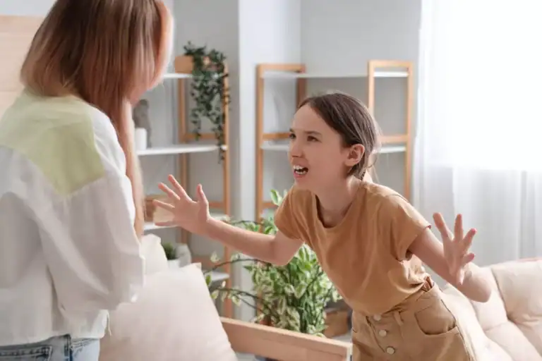 A little girl stands in front of her mom in a living room, raising both hands while arguing with her mom.