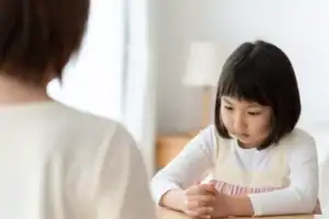 An adult seated across from a child at a table, with the child sitting upright and hands clasped, illustrating a scene associated with authoritarian parenting.