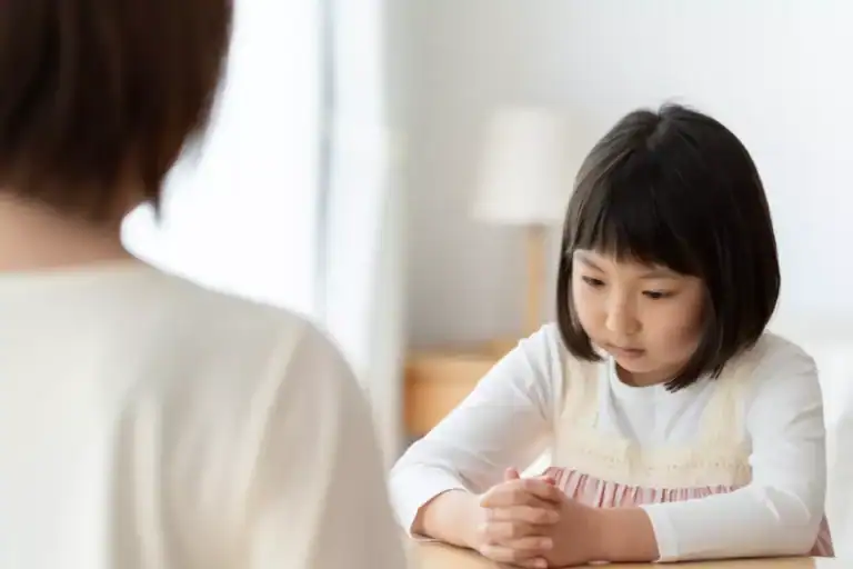 An adult seated across from a child at a table, with the child sitting upright and hands clasped, illustrating a scene associated with authoritarian parenting.