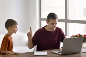 An adult seated at a desk points a finger upward while looking at a laptop, as a child stands nearby seeking attention, illustrating a scenario often associated with neglectful parenting.