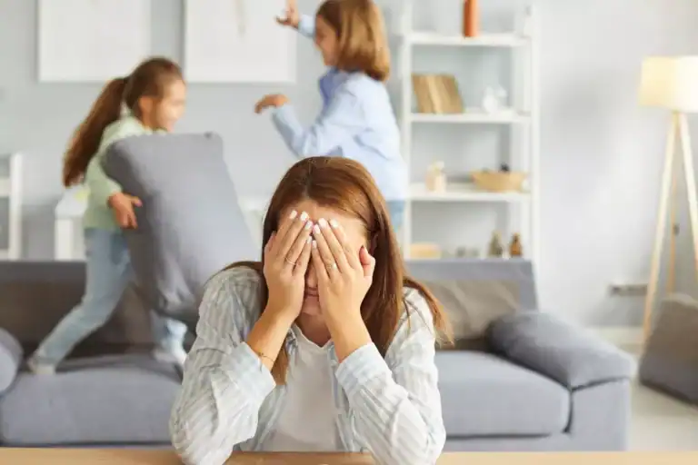 A mom sitting at a table with head resting in hands while two children play energetically on a couch behind them, illustrating a scene associated with permissive parenting.