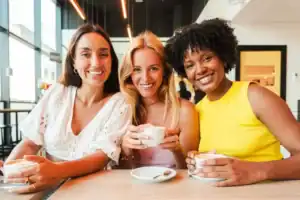 Three smiling women sit together at a café enjoying coffee and conversation in a bright, welcoming setting.