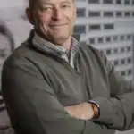 Drake Bassett standing with arms crossed, smiling, wearing a gray quarter-zip sweater over a plaid shirt, with a black-and-white urban building mural in the background.