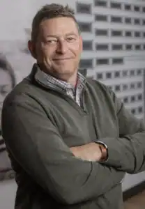 Drake Bassett standing with arms crossed, smiling, wearing a gray quarter-zip sweater over a plaid shirt, with a black-and-white urban building mural in the background.