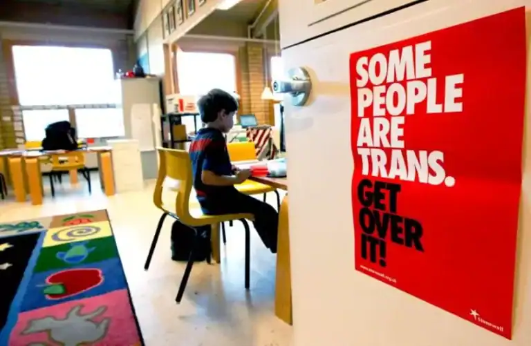 A young boy sitting in a school classroom by himself, with a sign on the door saying "Some People Are Trans. Get Over It!"