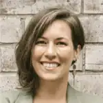 Headshot of Emma Fulenwider smiling in a sage green blazer against a brick wall background.