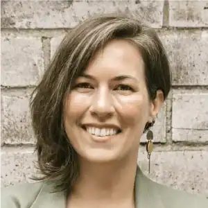 Headshot of Emma Fulenwider smiling in a sage green blazer against a brick wall background.