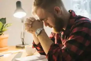 Young adult man praying with hands clasped over an open Bible at a desk, a moment of quiet holiness and reflection.