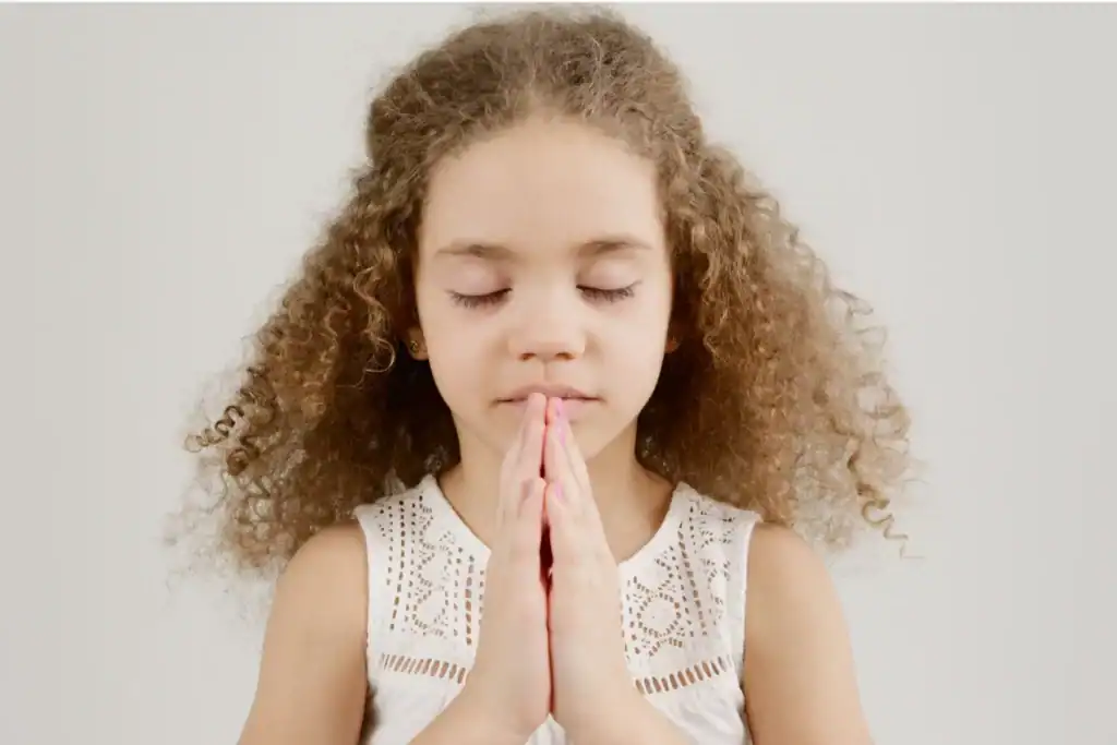 Young girl with curly hair stands with hands clasped in prayer, eyes closed, against a plain light background. This image emphasizes the importance of spiritual growth in young children.