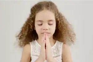 Young girl with curly hair stands with hands clasped in prayer, eyes closed, against a plain light background. This image emphasizes the importance of spiritual growth in young children.