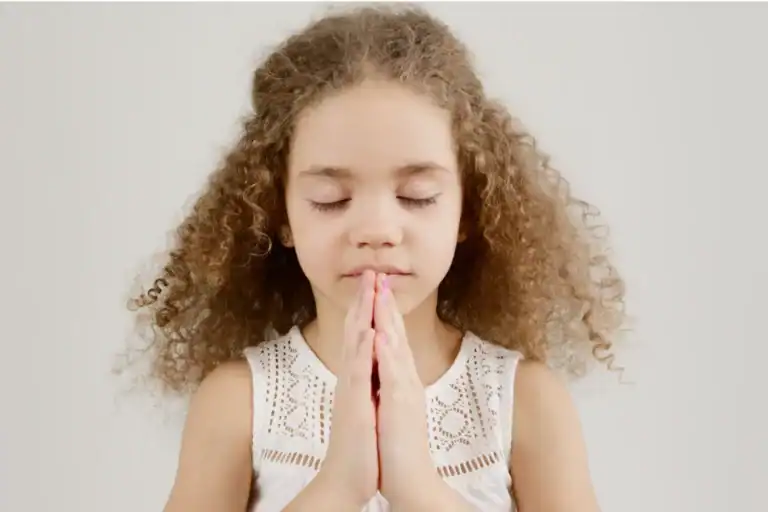 Young girl with curly hair stands with hands clasped in prayer, eyes closed, against a plain light background. This image emphasizes the importance of spiritual growth in young children.