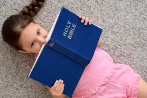 Little girl with brown hair lying on a carpeted floor while holding an open blue Bible in front of their face. The book cover displays the words “HOLY BIBLE” in gold lettering.