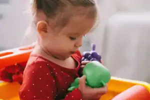 Toddler experiencing toy overload while holding oversized toy grapes and a green toy apple in a play seat.