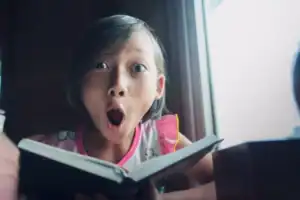 Little girl sitting by a window holding an open book, viewed close-up with wide eyes and an open mouth. Soft natural light from the window illuminates the face, while stacks of books appear blurred in the foreground.
